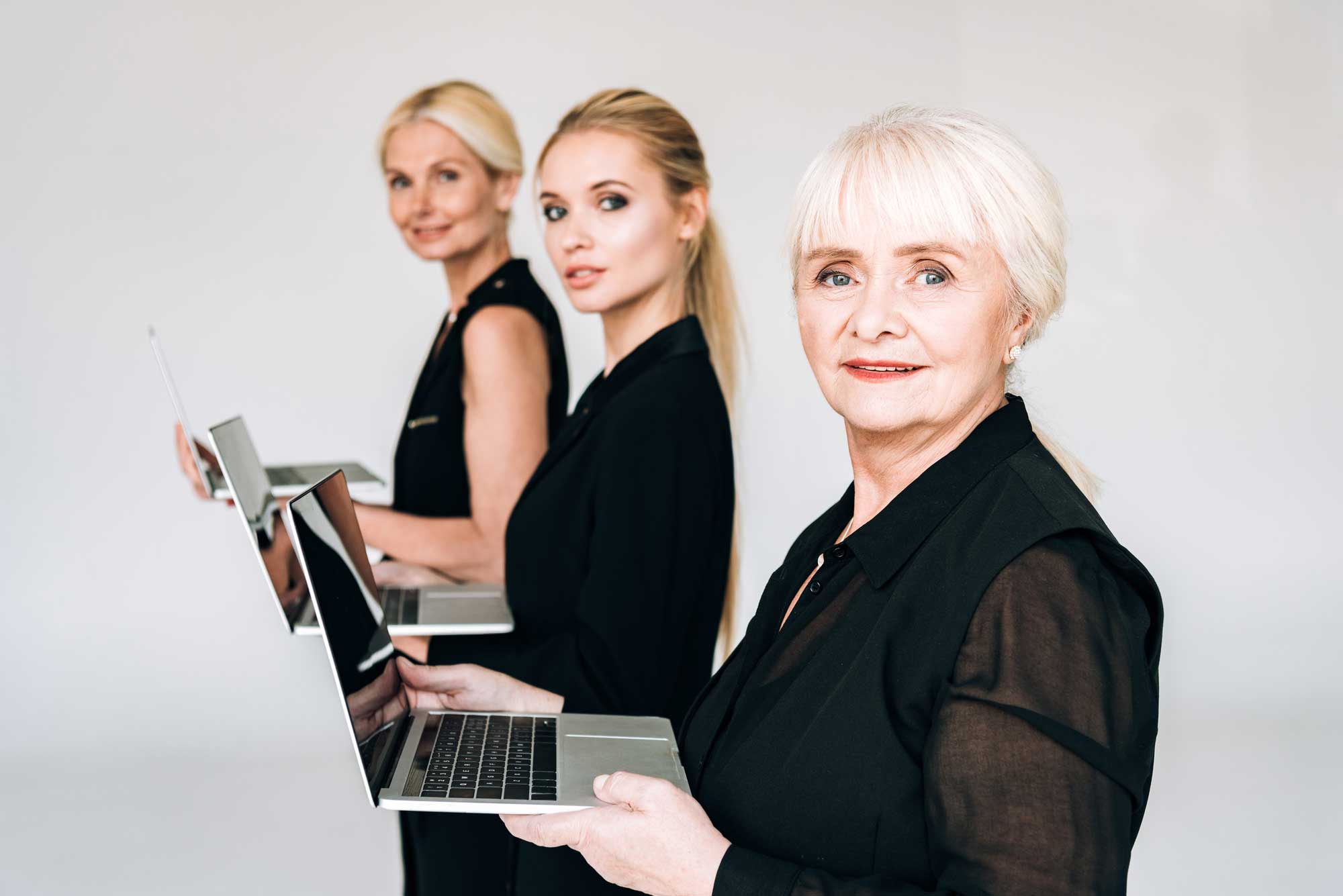 Three blonde women of different generations stand in a row holding laptops, looking over their left shoulders toward the camera.