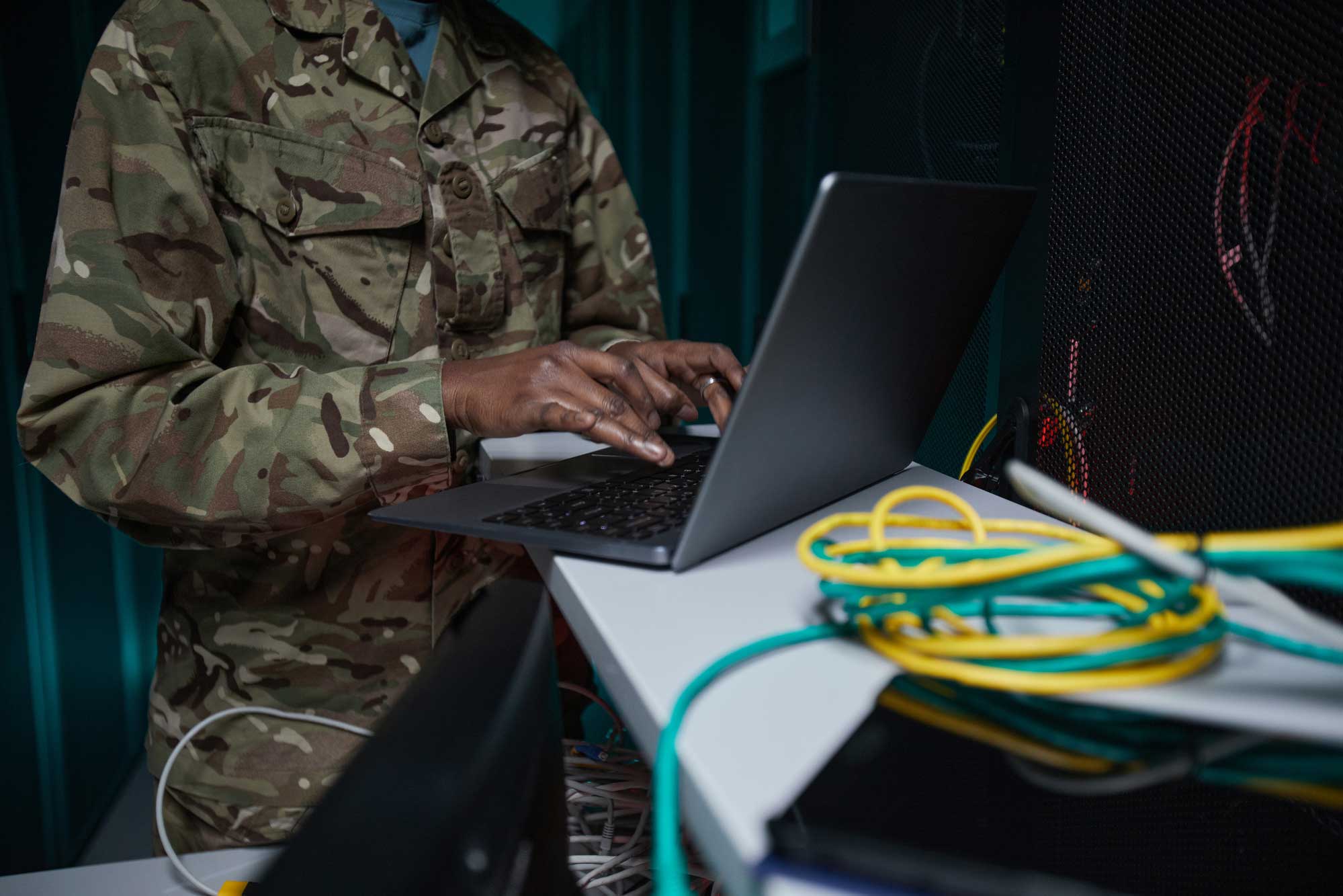A person in a military uniform uses a laptop to set up a network in a server room.